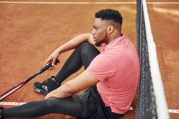 Fototapeta na wymiar Takes a break. African american man in pink shirt plays tennis on the court outdoors