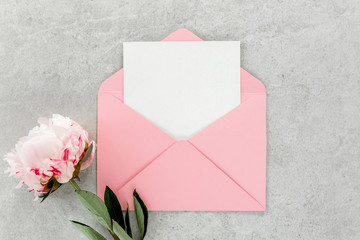 Mockup invitation, blank paper greeting card, pink envelope and peonies on gray stone table. Flower background. Flat lay, top view.