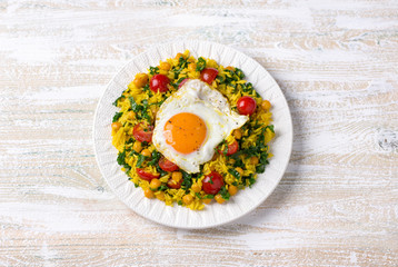 Chinese fried rice with turmeric, chickpeas, tomatoes, kale and with fried eggs on white ceramic plate on wooden background