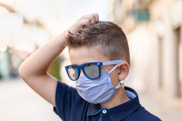 A boy in sunglasses and a medical mask scratches his head in the street