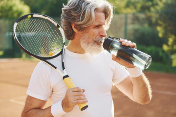 Taking a break and drinking water. Senior modern stylish man with racket outdoors on tennis court at daytime