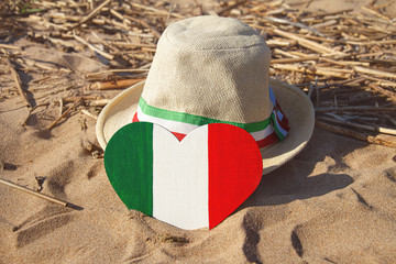 wooden heart and straw hat with ribbon with colors of flag of Italy green, white and red in golden sand on beach