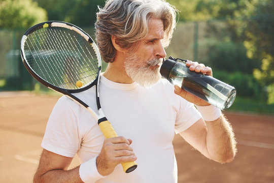 Taking a break and drinking water. Senior modern stylish man with racket outdoors on tennis court at daytime