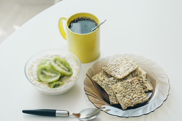 healthy breakfast on a white table in natural sunlight. Oatmeal with exotic fruits, whole grain bread, a cup of coffee. High quality photo