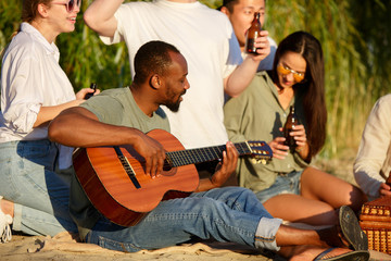 Guitar playing. Group of friends clinking beer glasses during picnic at the beach in sunshine. Lifestyle, friendship, having fun, weekend and resting concept. Looks cheerful, happy, celebrating