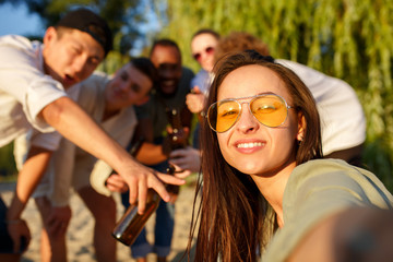 Evening. Group of friends clinking beer glasses during picnic at the beach in sunshine. Lifestyle, friendship, having fun, weekend and resting concept. Looks cheerful, happy, celebrating, festive.