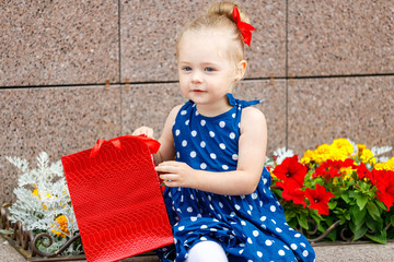 a little girl in a blue dress and a red bow sits with colored bags on the street next to flowers