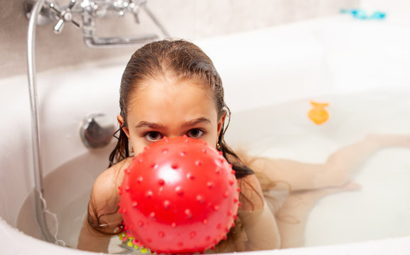 Cheerful little smiling charming girl bathes with red ball and looks at the camera. The concept of entertainment at home for children during quarantine