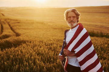 View from behind. Holding USA flag in hands. Patriotic senior stylish man with grey hair and beard on the agricultural field