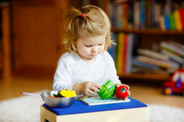 Cute little toddler girl playing at home with eco wooden toys. Happy healthy excited child cutting vegetables and fruits with toy knife. Baby girl having fun with role kitchen and cooking game.