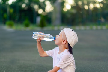 portrait of a blond boy in a sports uniform sitting on a green lawn on a football field and drinking water from a bottle, sports section. Training of children, children's leisure