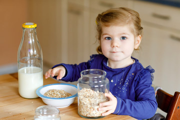 Adorable toddler girl eating healthy oatmeals with milk for breakfast. Cute happy baby child in...