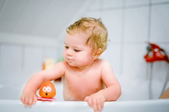 Cute adorable baby girl taking foamy bath in bathtub. Toddler playing with bath rubber toys. Beautiful child having fun with colorful gum toys and foam bubbles