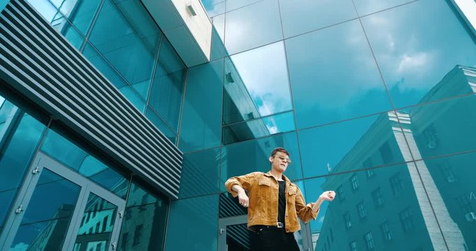 Happy young boy having fun outdoor in street wearing glasses, celebrating youth, active lifestyle, party maker. Stylish dancer man moves comically rhythmically against background of glass building