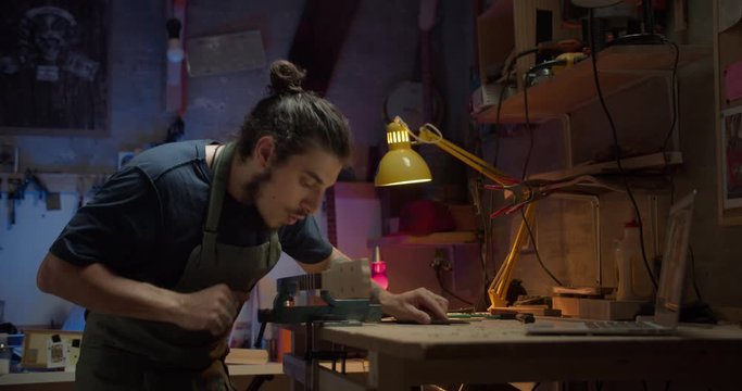 Attractive brunette luthier blowing out wood dust from musical instrument guitar arm, young joiner in apron working on wooden table using carpenter tools instruments, blue and red light on background