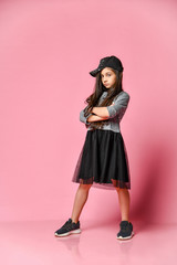 Portrait of a little hipster girl in a dark dress with a baseball cap and sneakers, on a pink...