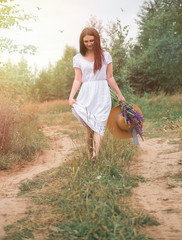 Beautiful girl on a village road in a white dress with wild flowers and a summer hat barefoot