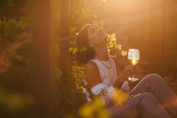 Young beautiful woman drinking wine in vineyard at sunset on summer sunny day.