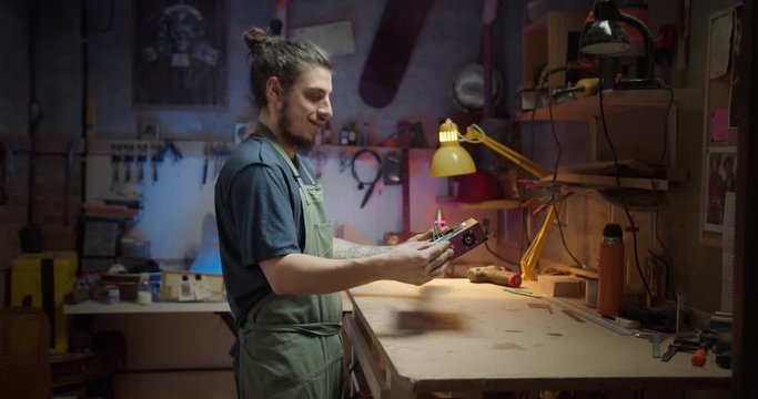 Side shot of happy master luthier result of work project musical instrument created indoor, evening studio blue and red light, hobby and art concept in workshop of Hispanic male carpenter with desk