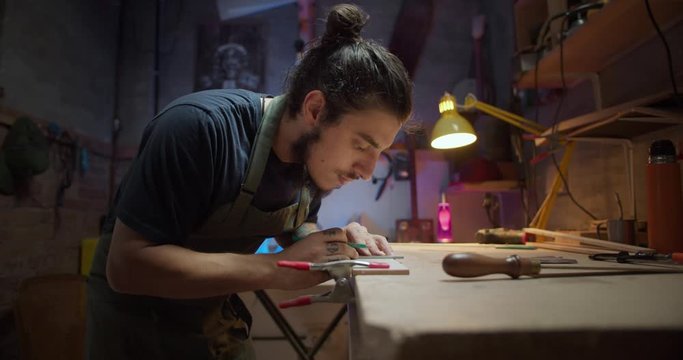 Close up portrait of work process of professional carpenter artisan master in cozy studio, handsome young joiner handcraft at workshop desk with light colorful lamp marking using pencil and tools