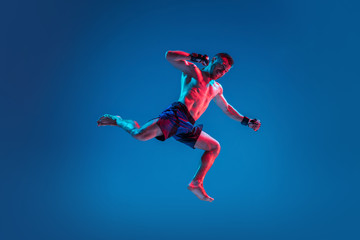 Naklejka na ściany i meble High flight. MMA. Professional fighter punching or boxing isolated on blue studio background in neon. Fit muscular caucasian athlete or boxer fighting. Sport, competition and human emotions, ad.
