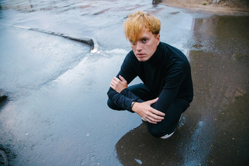 A guy in black sits on his haunches on the wet asphalt and looks at the viewer.
