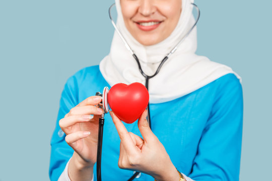 Portrait Of A Friendly, Muslim Woman Doctor Or Nurse With A Stethoscope And A Red Heart In Her Hand. On A Blue Background.