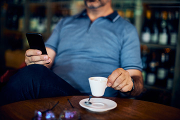 Mature man sitting in coffee shop, drinking coffee and using smart phone.