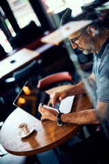 Mature man sitting in coffee shop and using smart phone. There is a coffee and laptop on desk.