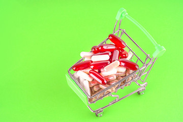 Multicolored capsules with biologically active additives and vitamins in the shopping cart on a green background with copy space. Top view. Preventive and alternative medicine. 
