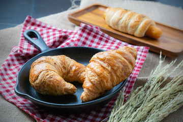 Two pieces of croissant on black ceramic pan that put on the fabric near rice in paddy with sausage bread behind them in yellow sun light for morning meal or breakfast for carbohydrate source.