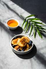 Mini Pancakes mit frischen Heidelbeeren in einer Schüssel und Honig auf einem grauen Hintergrund. Licht, Schlagschatten, Kontrast, Frühstück.