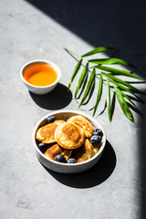 Mini Pancakes mit frischen Heidelbeeren in einer Schüssel und Honig auf einem grauen Hintergrund. Licht, Schlagschatten, Kontrast, Frühstück.
