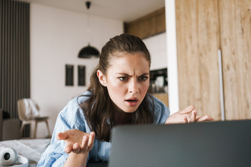 Photo of displeased caucasian girl using laptop while lying on bed