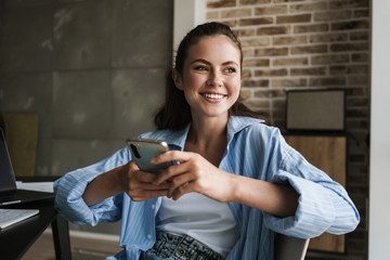 Fototapeta premium Photo of joyful nice girl smiling and using smartphone while sitting