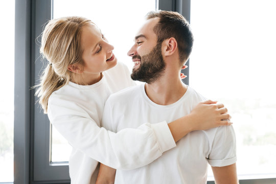 Image of happy couple smiling and hugging while standing near window