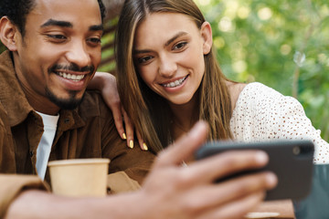 Image of multinational couple using mobile phone while drinking coffee