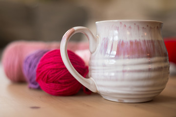 Cozy mug next to yarn balls ready for knitting on a light colored wood coffee table.  Gray sectional sofa in background.  Light pink, purple, and red yarn just behind large mug with 4 finger handle.