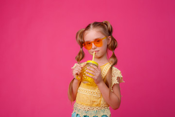 portrait of a little blonde girl in sunglasses on pink with a glass jar of cooling cocktail.