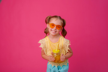 portrait of a little blonde girl in sunglasses on pink with a glass jar of cooling cocktail.