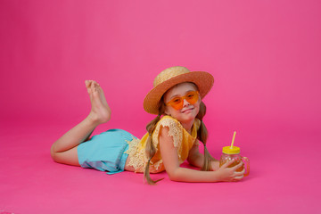 Obraz na płótnie Canvas a little girl in sunglasses , a straw hat and a cocktail is lying on a pink background