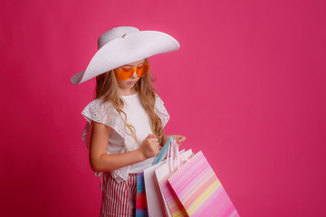 a little Girl with a lot of shopping bags, wearing sunglasses and a hat smiles on a pink background in the Studio, shopping concept, sale