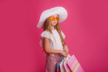 a little Girl with a lot of shopping bags, wearing sunglasses and a hat smiles on a pink background in the Studio, shopping concept, sale