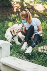 Front view photo of a blonde woman wearing an anti virus mask lying on the grass with her labrador