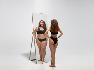 Young fit, slim woman looking at fat girl in mirror's reflection on white background. Thinking...