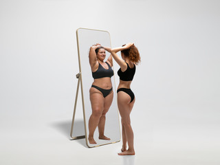Young fit, slim woman looking at fat girl in mirror's reflection on white background. Thinking...