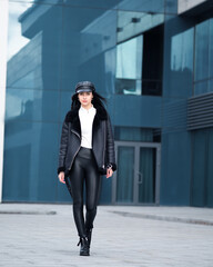 Young brunette woman in the leather pants sheepskin coat and black cappi posing at the city