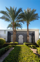 Obraz premium High Palm Trees And Old Wooden Arch In Egypt. Middle East Architecture And Ornamental Garden In Resort City, Green Bushes And Paved Path. Tourist Place.