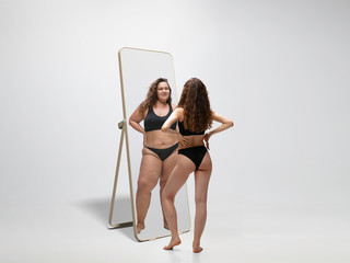 Young fit, slim woman looking at fat girl in mirror's reflection on white background. Thinking...