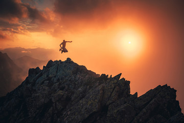 Happy man in mountains. Young hiker jumping on the top of the hill, sunset sky in background,..